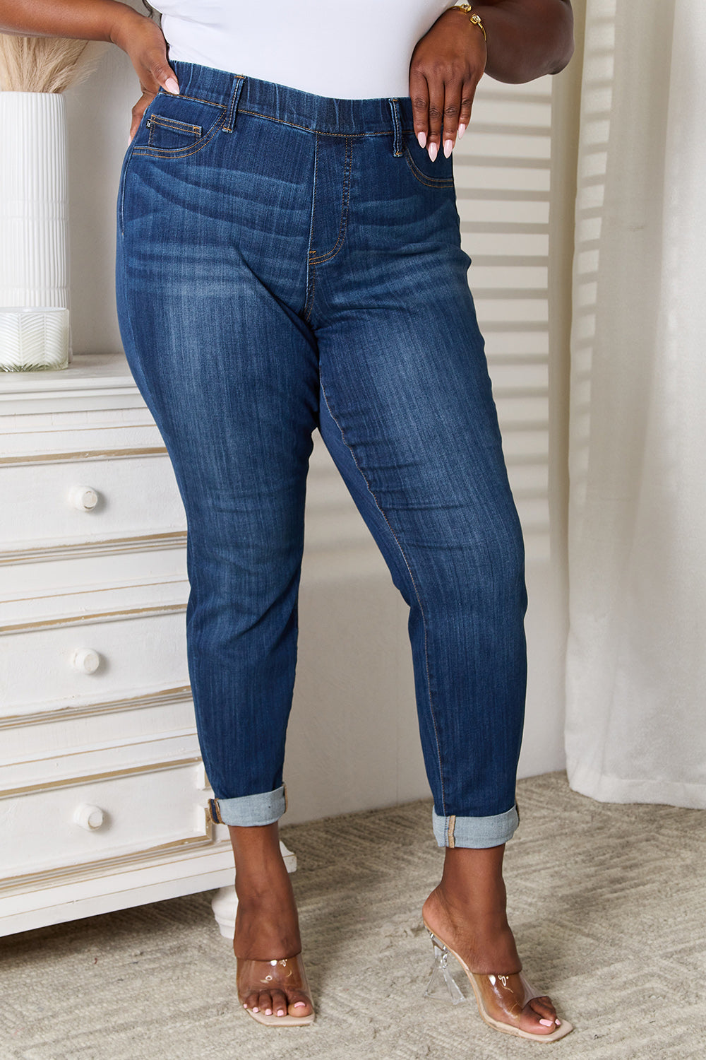 A person wearing Judy Blue Full Size Skinny Cropped Jeans with rolled-up cuffs and clear heels stands near a white dresser adorned with a decorative vase for a modern look.