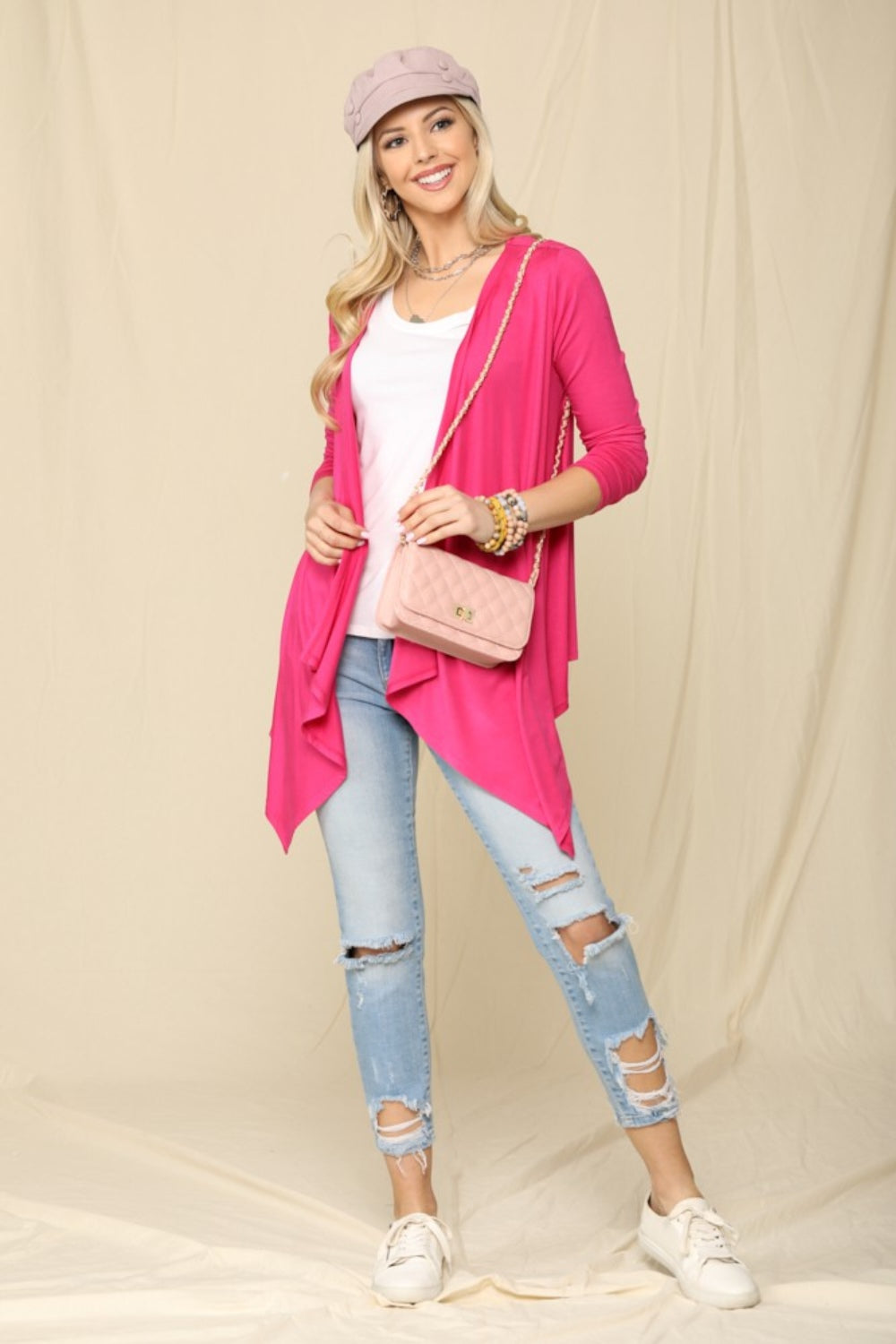 A woman dressed in the Celeste Full Size Open Front Knit Cardigan in soft pink, layered over a white top. Her ripped jeans add an edgy touch, while the light pink cap and matching handbag complete the look as a versatile wardrobe staple.