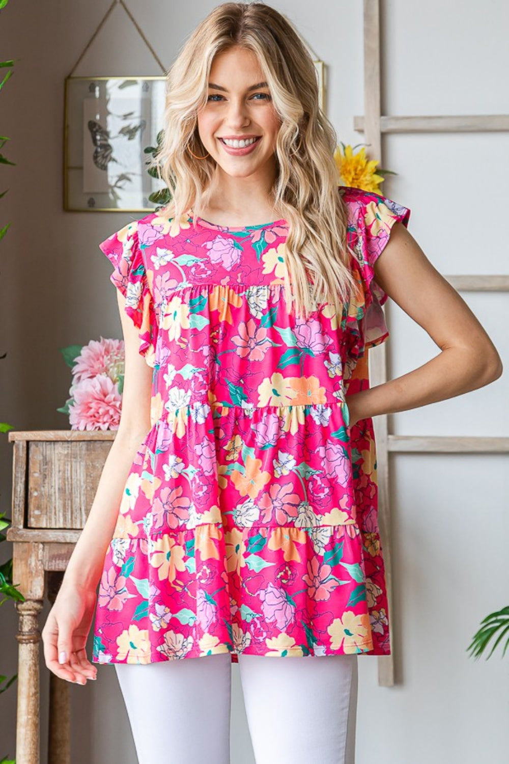 A woman with long blonde hair wearing a Heimish Full Size Floral Ruffle Sleeve Tiered Blouse and white pants smiles while standing in a room with plants and decor.