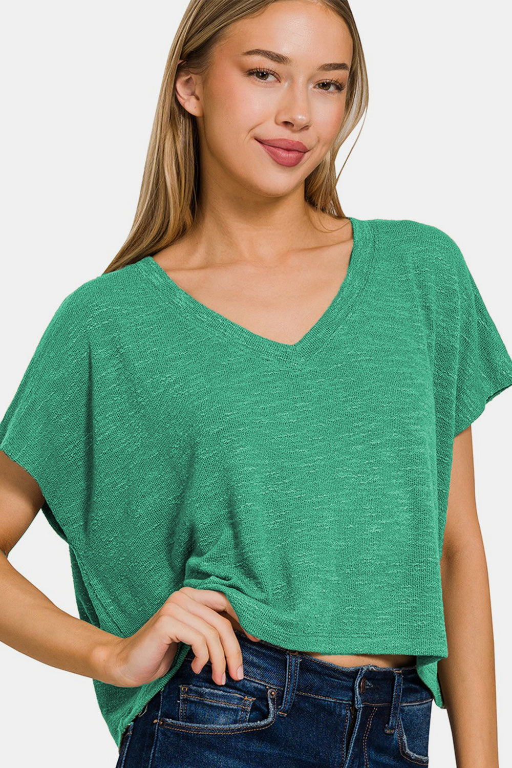 A person with long hair wears a green Zenana V-Neck Short Sleeve T-Shirt and blue jeans, standing against a white background. This versatile wardrobe essential offers both a stylish look and comfortable fit.