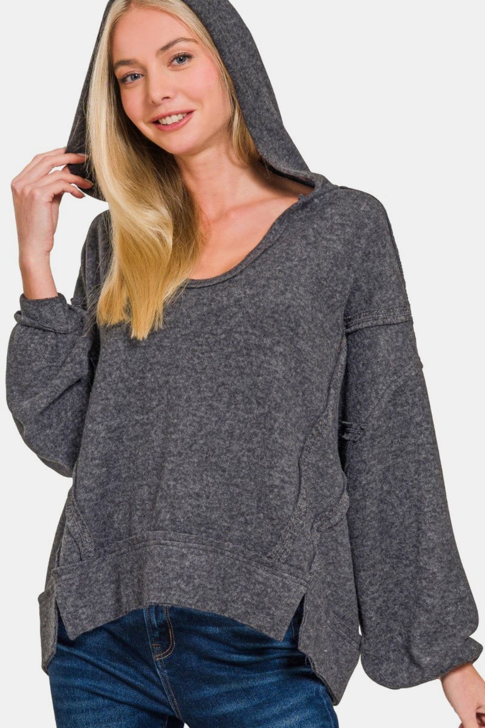 A person with long blond hair stands against a white background, wearing the Zenana Brushed Hacci Exposed Seam Hoodie in dark gray paired with jeans.