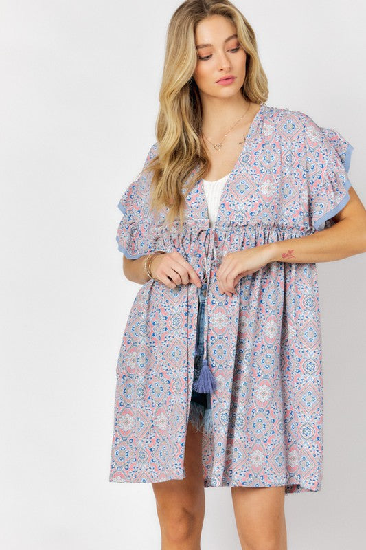 A woman stands against a white background wearing the Printed Short Sleeve Ruffle Kimono over a white top and denim shorts, effortlessly blending style and comfort.