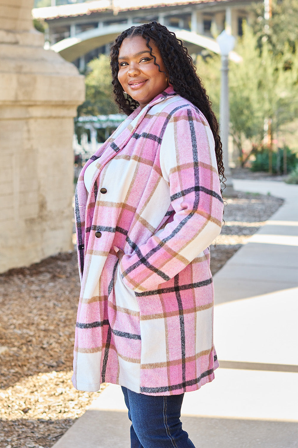 A woman stands on a sidewalk wearing the Double Take Full Size Plaid Button Up Lapel Collar Coat in pink and white over a white top and blue jeans, with her right hand lightly touching her hair. The polyester fabric of the coat makes it durable and perfect for an easy machine wash cold.