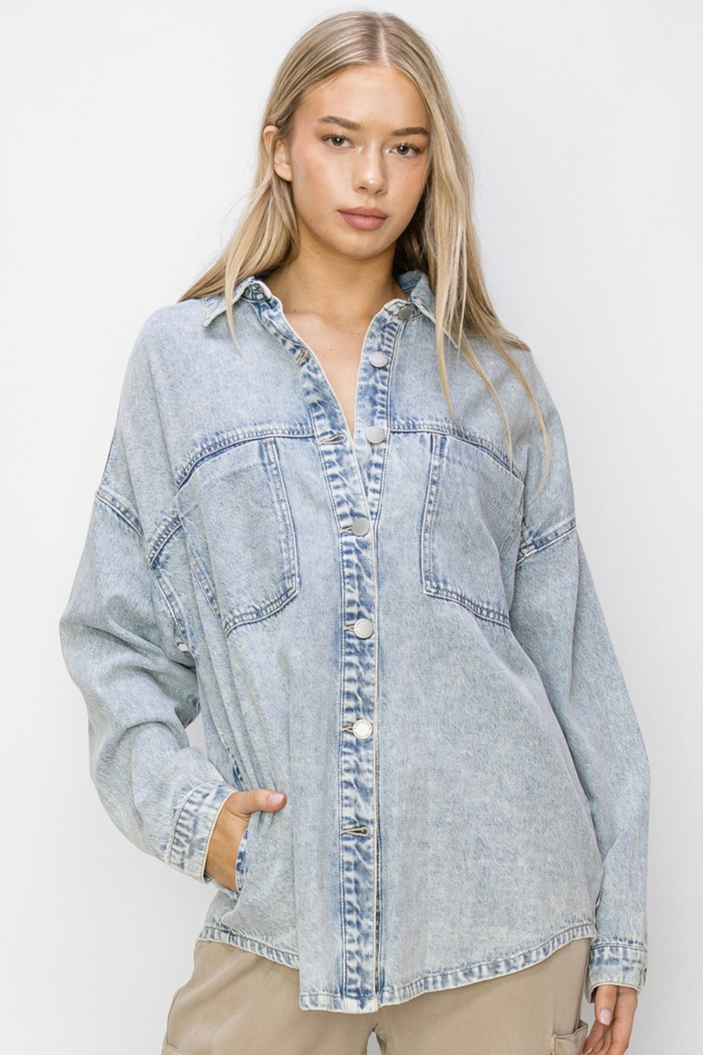 A person with long hair wearing a RISEN Button Up Long Sleeve Shacket in a light-wash denim stands against a plain background with a neutral expression.