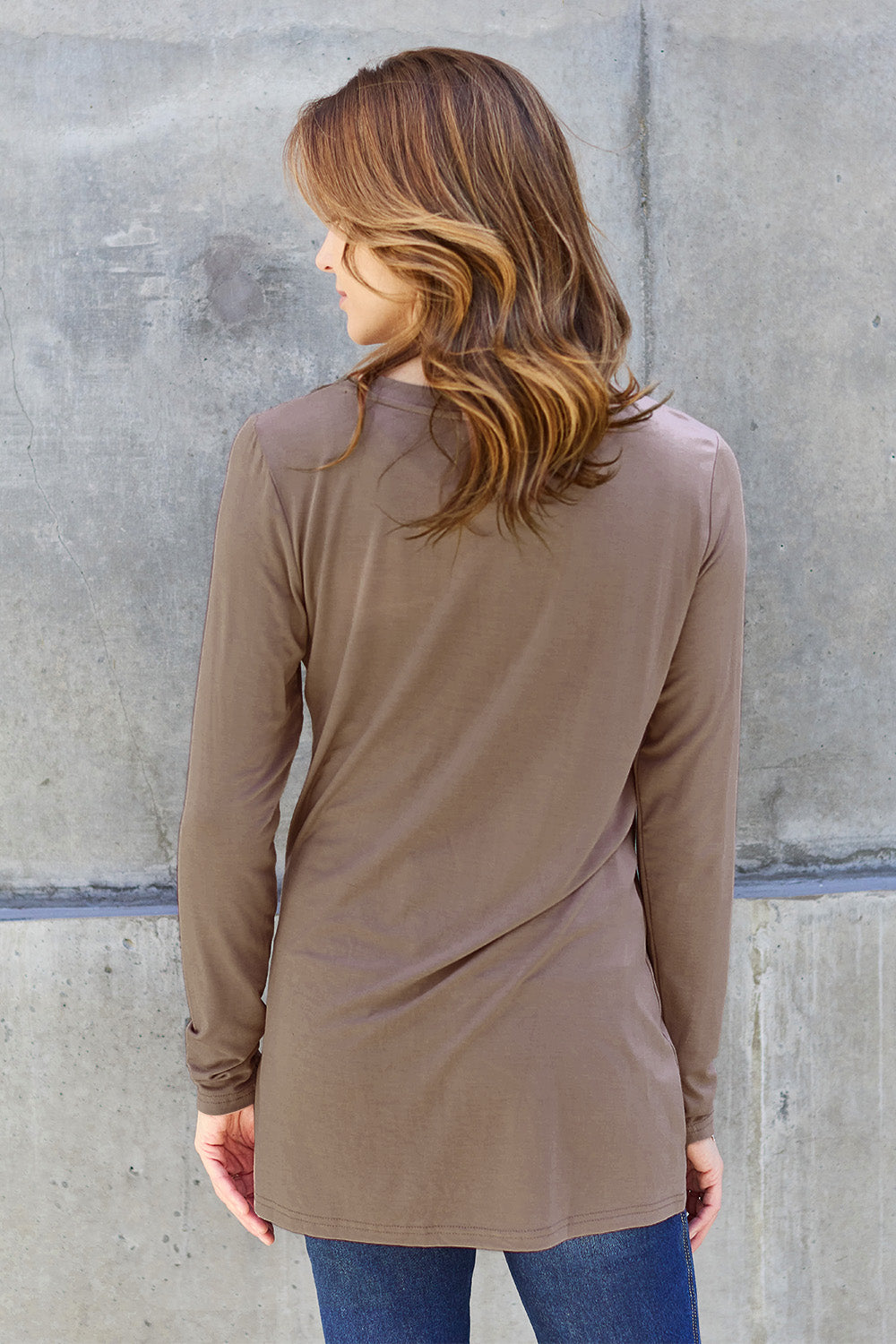 A woman with long hair wearing sunglasses, a Basic Bae Full Size Round Neck Long Sleeve Top in turquoise, and jeans is standing in front of a concrete wall with one hand on her hip.