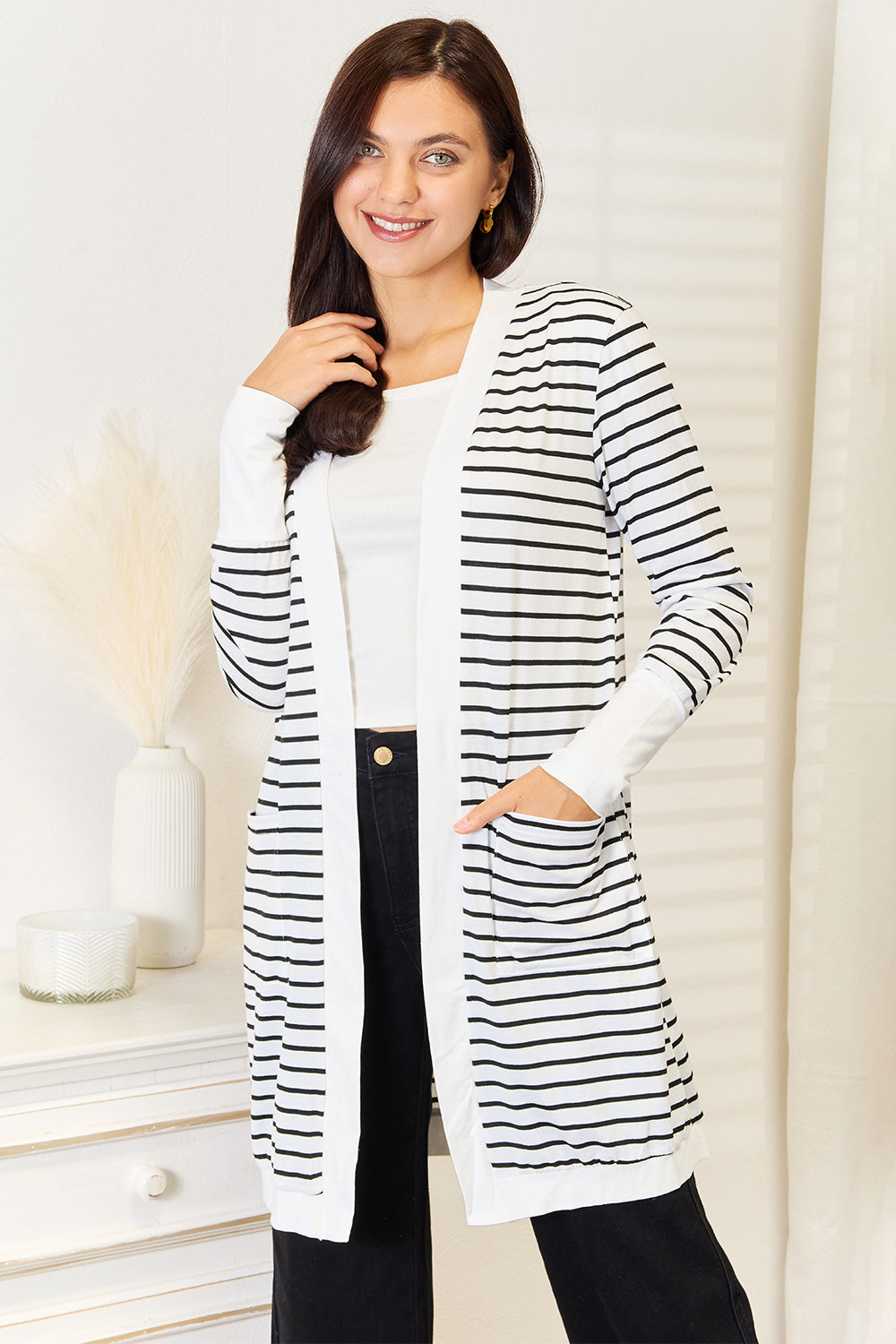 A woman wearing the Double Take Striped Open Front Longline Cardigan over a white top and black pants stands indoors, smiling at the camera. She is posing with one hand on her hip, showcasing this versatile piece.