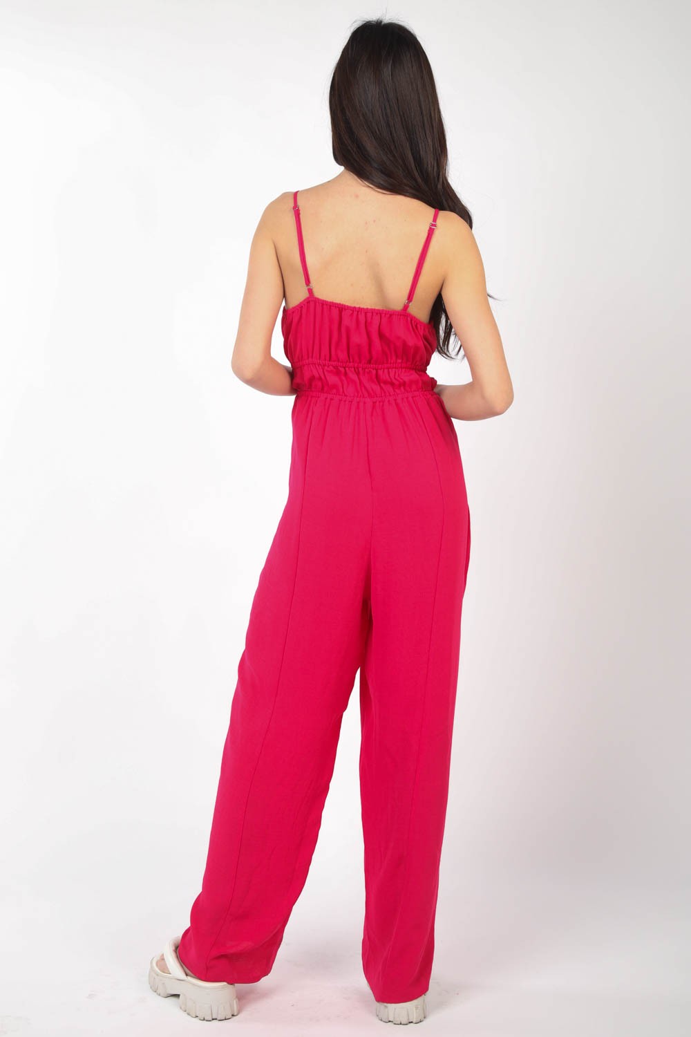 A person wearing the VERY J Pintuck Detail Woven Sleeveless Jumpsuit in bright pink paired with white sandals stands against a white background, with one hand placed in a pocket.