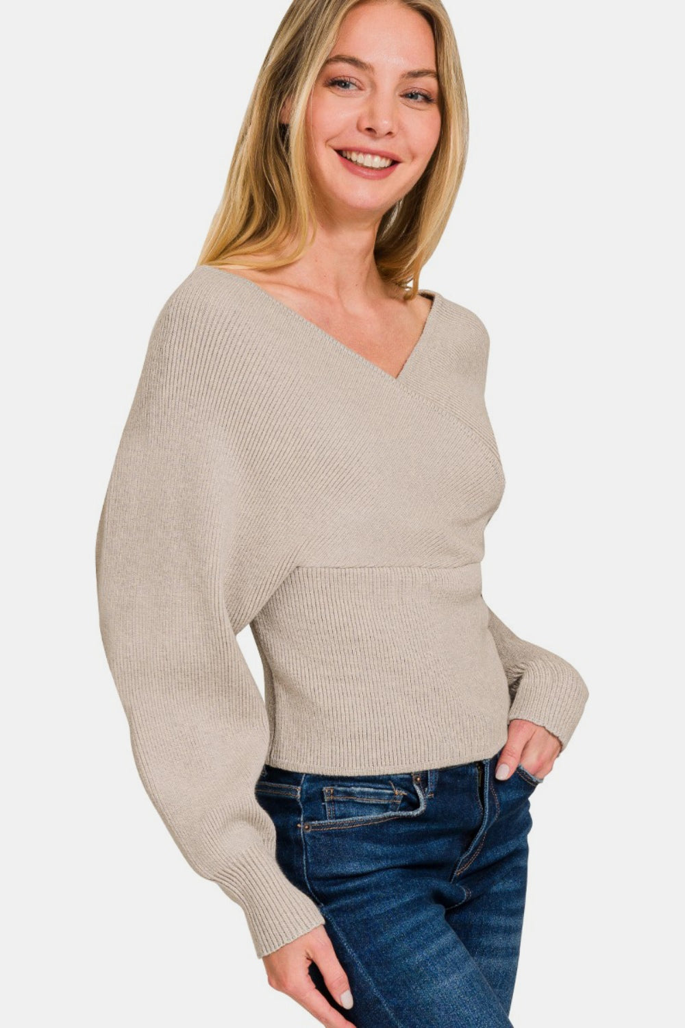 A woman with long blonde hair is smiling with one hand in her pocket against a plain white background, wearing the Zenana Cross Wrap Rib Long Sleeve Sweater in beige and blue jeans—a perfect layering option for any season.