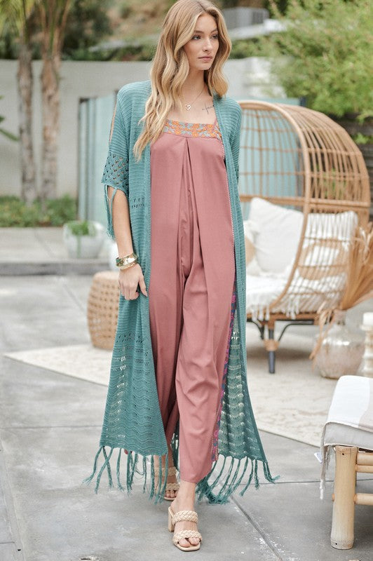 A woman in a green floral dress and a Solid Long Cardigan With Fringe exudes boho-chic vibes as she strolls on a patio, surrounded by outdoor furniture and lush plants.