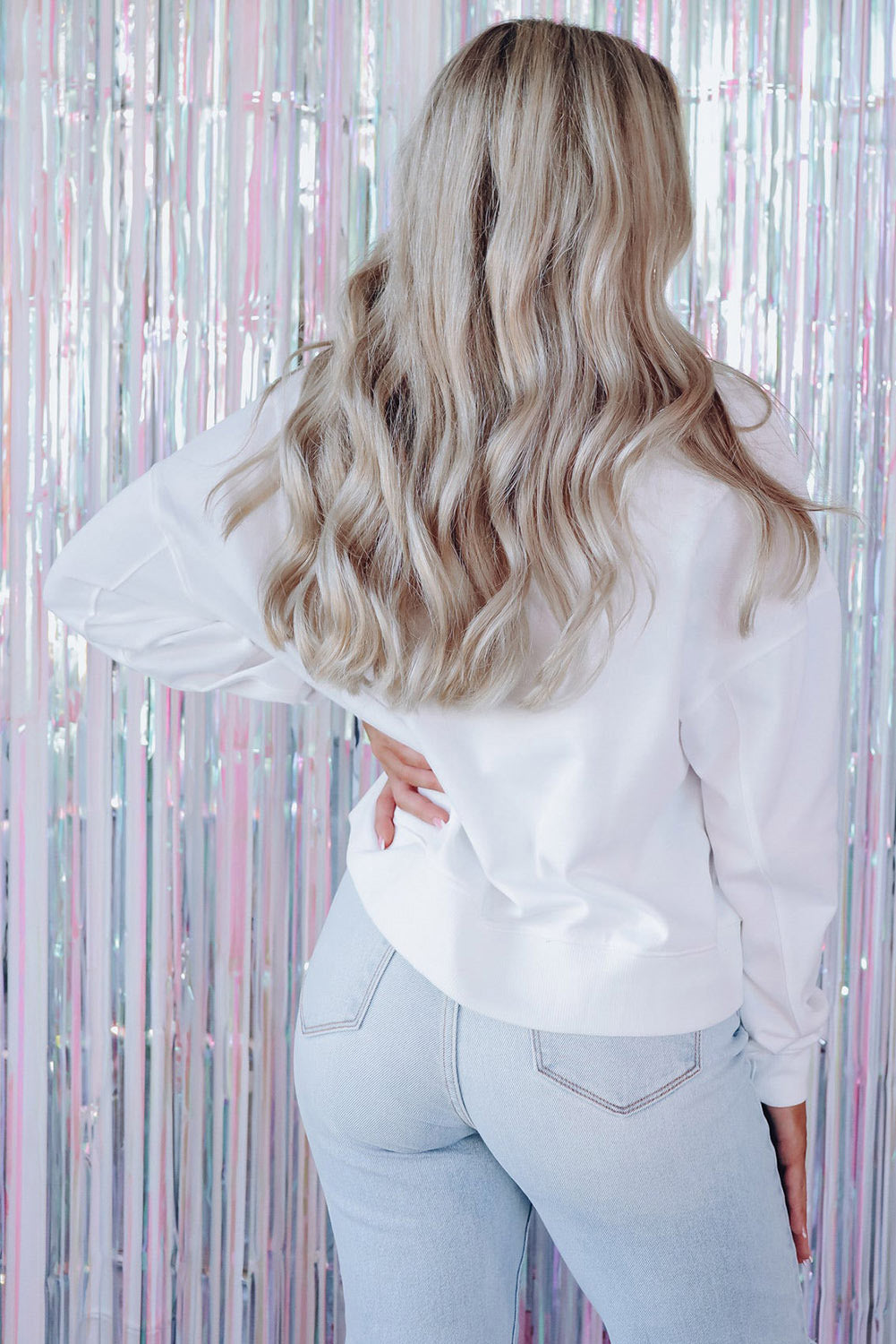 A person with long blonde hair is standing with their back to the camera, wearing a White BEACH BABE Slogan Graphic Casual Sweatshirt and light blue jeans.