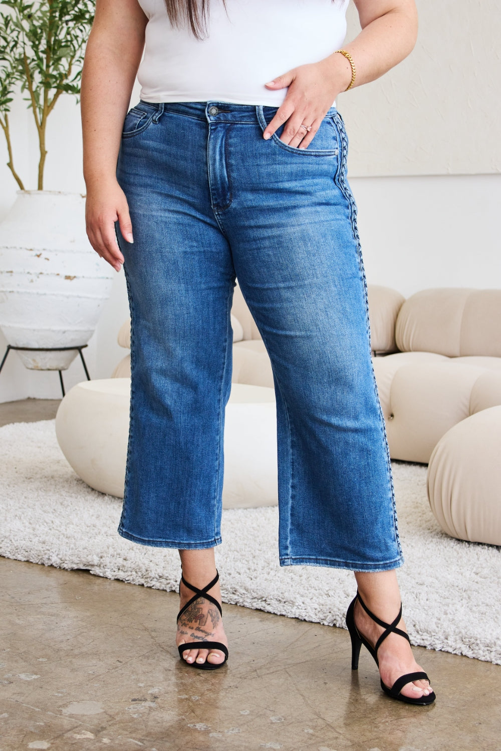 A person wearing Judy Blue Full Size Braid Side Detail Wide Leg Jeans and black heels is standing on a polished floor. There is a white couch and a large potted plant in the background.