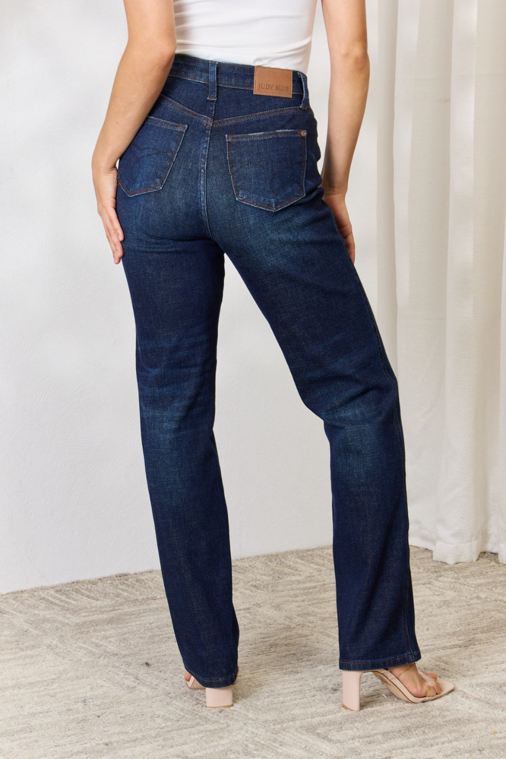 A person stands with hands in pockets, wearing the Judy Blue Full Size Button-Fly Straight Jeans, known for their high-waisted dark blue design crafted from premium lycra fabric. The jeans showcase button details and a straight leg cut, complemented by a white top and open-toed heels.