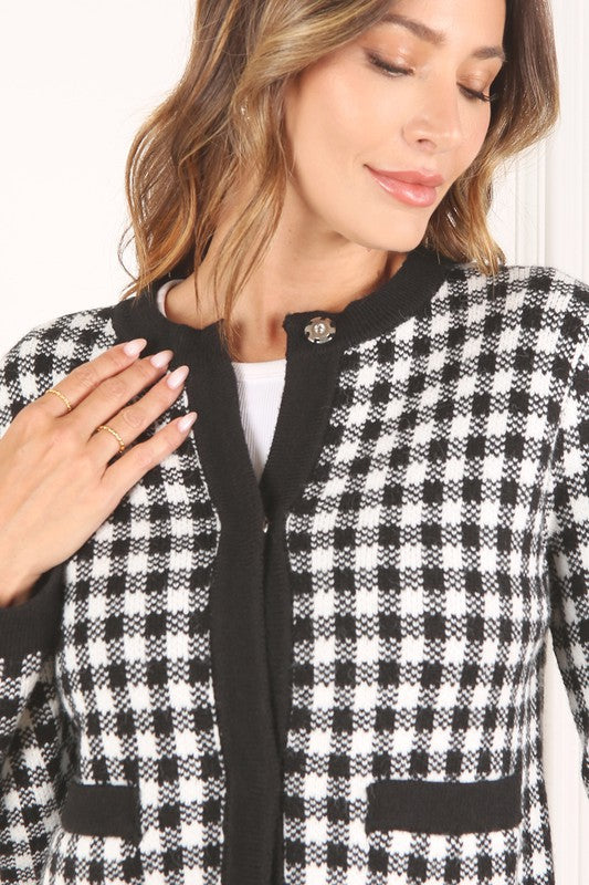 A woman dressed in the Black Check Knitted Jacket, featuring snap buttons and functional pockets, complemented by black pants, stands against a white background.