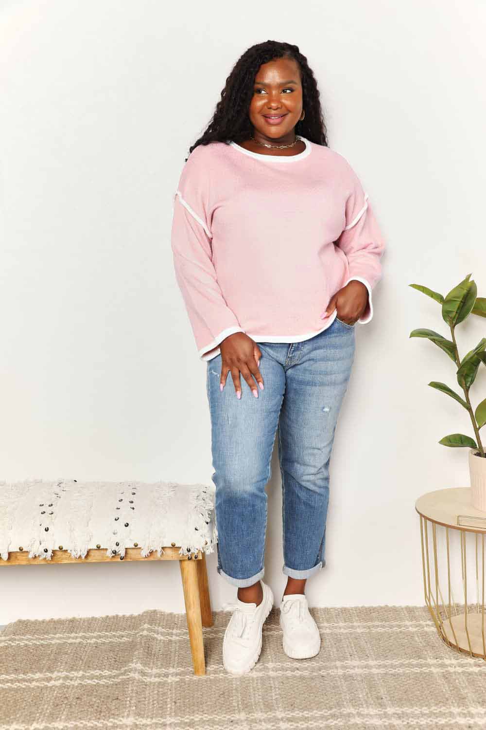 A woman with long curly hair is wearing the Angel Wings Contrast Detail Dropped Shoulder Knit Top in pink, paired with blue jeans, standing against a light background.