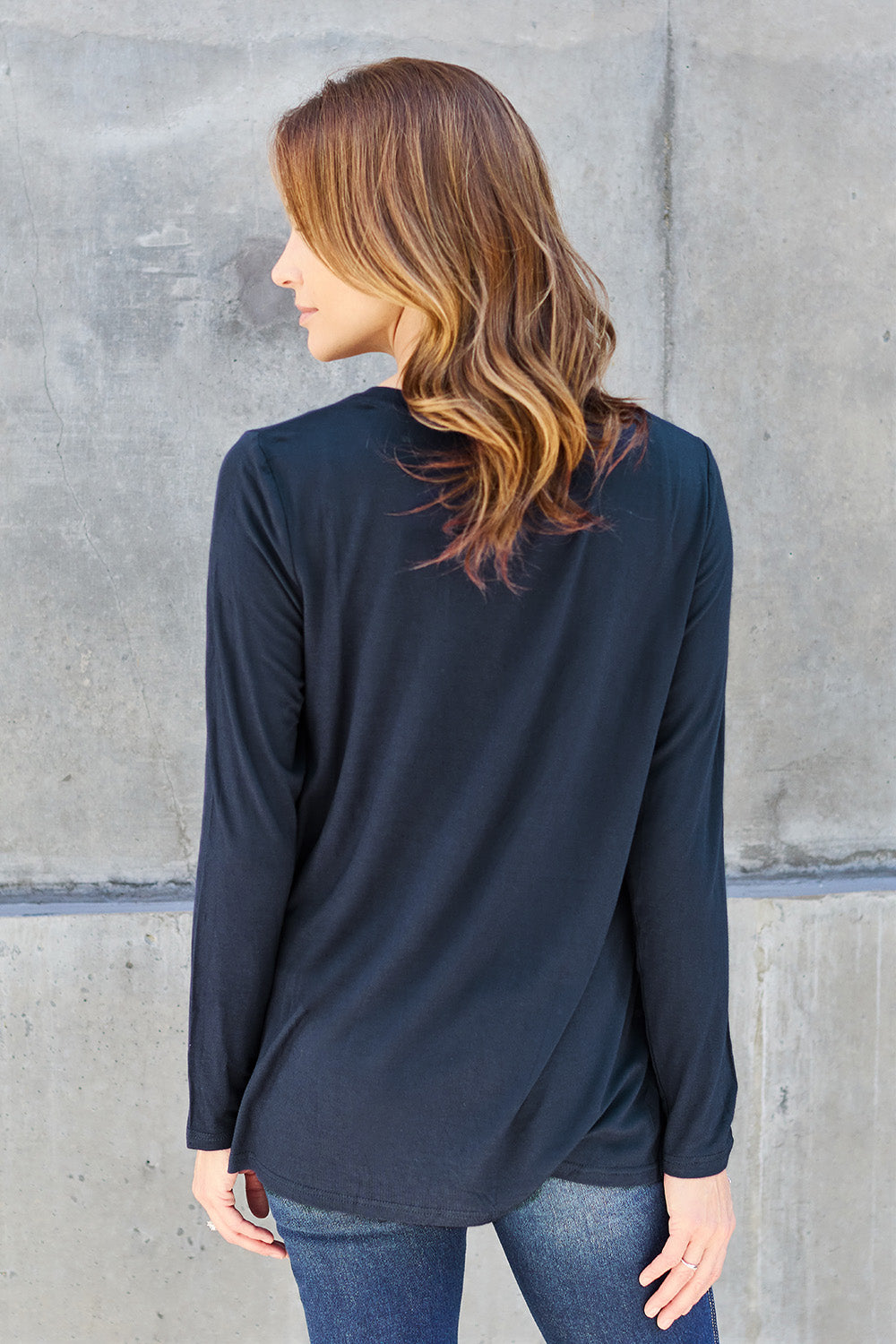 A woman with light brown hair stands against a concrete wall, wearing the Basic Bae Full Size V-Neck Long Sleeve Top in olive green and blue jeans, with a white backpack on her shoulder. Her outfit exudes basic style and is made from polyester, ensuring it’s both comfortable and suitable for machine wash cold. She is looking at the camera.