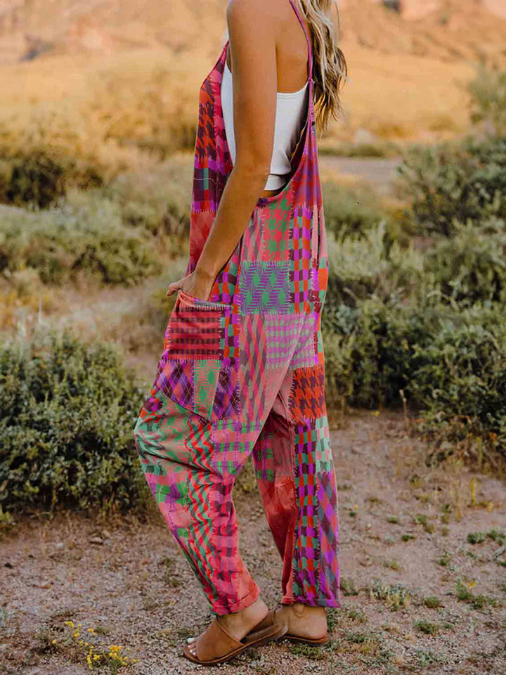 A woman stands outdoors in front of a rocky landscape, wearing the Full Size Printed V-Neck Sleeveless Jumpsuit and a brown brimmed hat. She is smiling and holding the brim of her hat with one hand, exuding the carefree vibe of this vibrant and colorful jumpsuit.