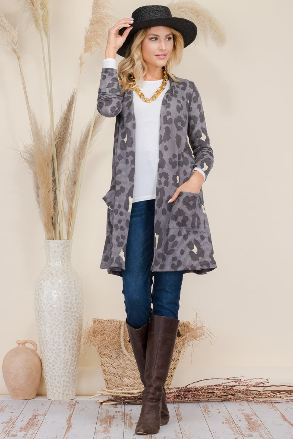 A person wearing a Celeste Full Size Leopard Open Front Contrast Cardigan over a white top and jeans, complemented by brown boots, accessorized with a black hat and necklace, stands next to a tall vase with decorative grasses.