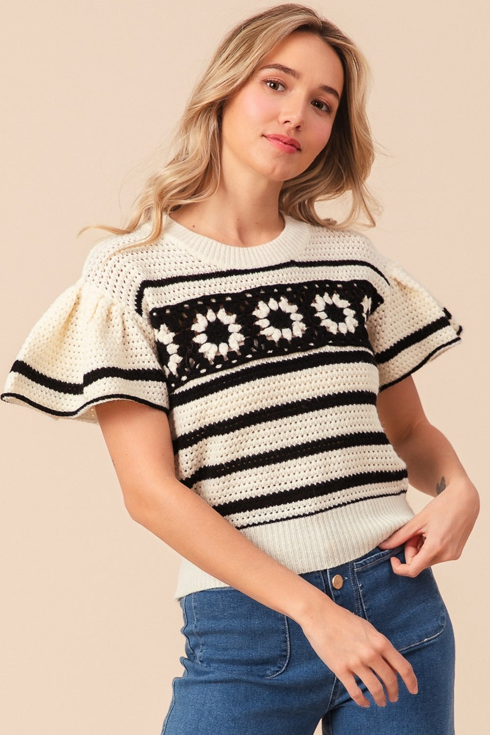 A woman sporting the BiBi Granny Square Short Sleeve Striped Sweater, which features cream and black stripes with floral crochet patterns on the chest, paired with blue jeans. She has light hair and is posing against a neutral background.