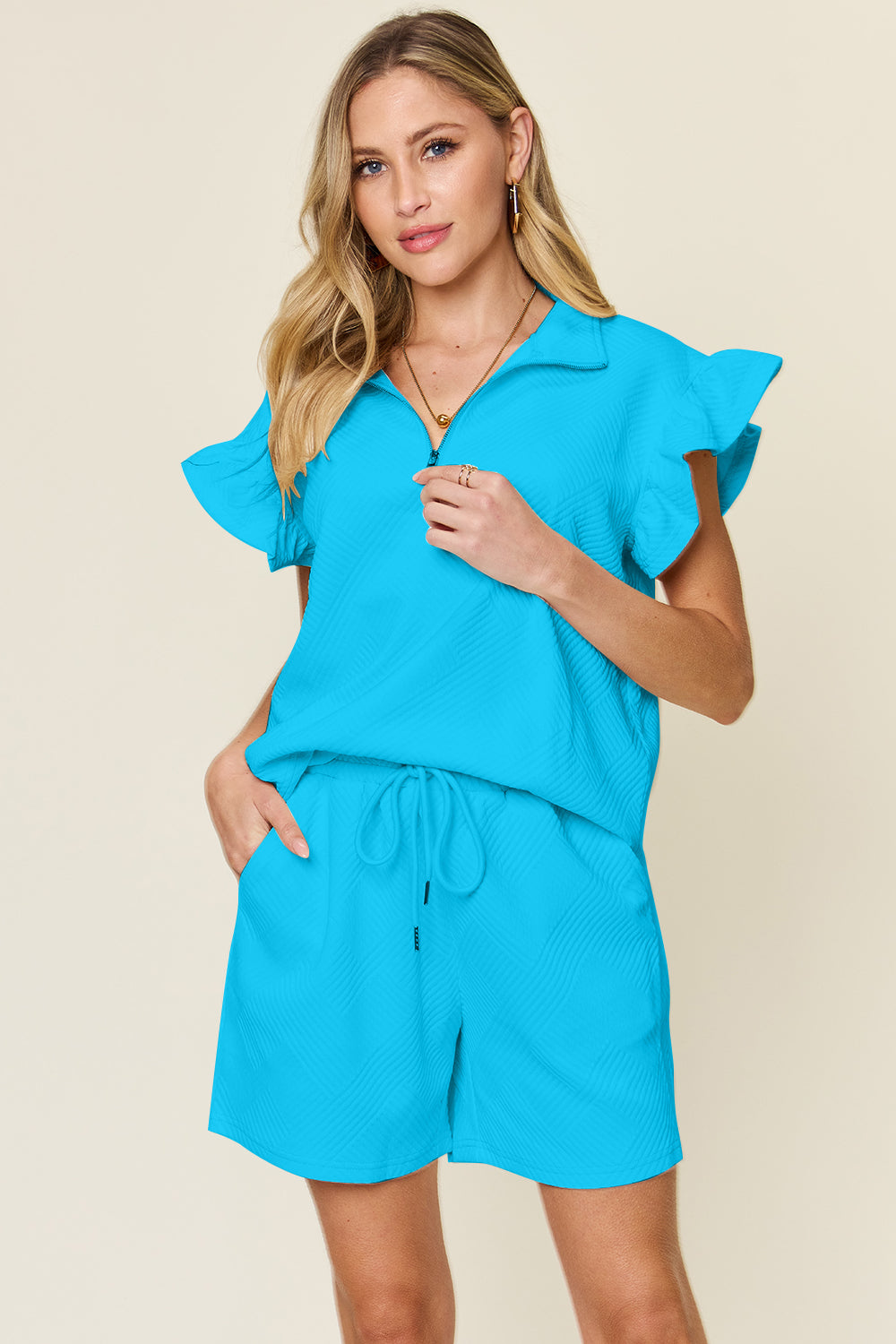 Against a plain background, a woman with blonde hair poses confidently with one hand in her pocket, highlighting the chic style of her Double Take Full Size Texture Flounce Sleeve Top and Drawstring Shorts Set.