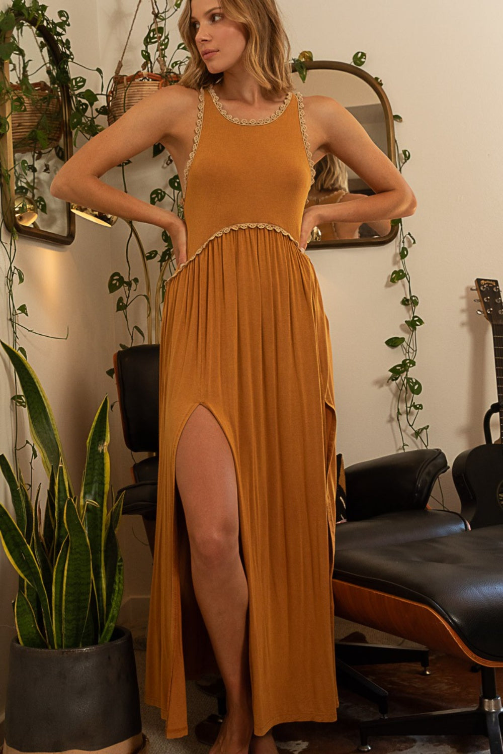 A woman stands barefoot in a room wearing a POL Sleeveless Back Zipper Front Slit Maxi Dress in mustard yellow, leaning against a doorframe. The versatile dress adds to her wardrobe, complemented by plants and a chair in the background.