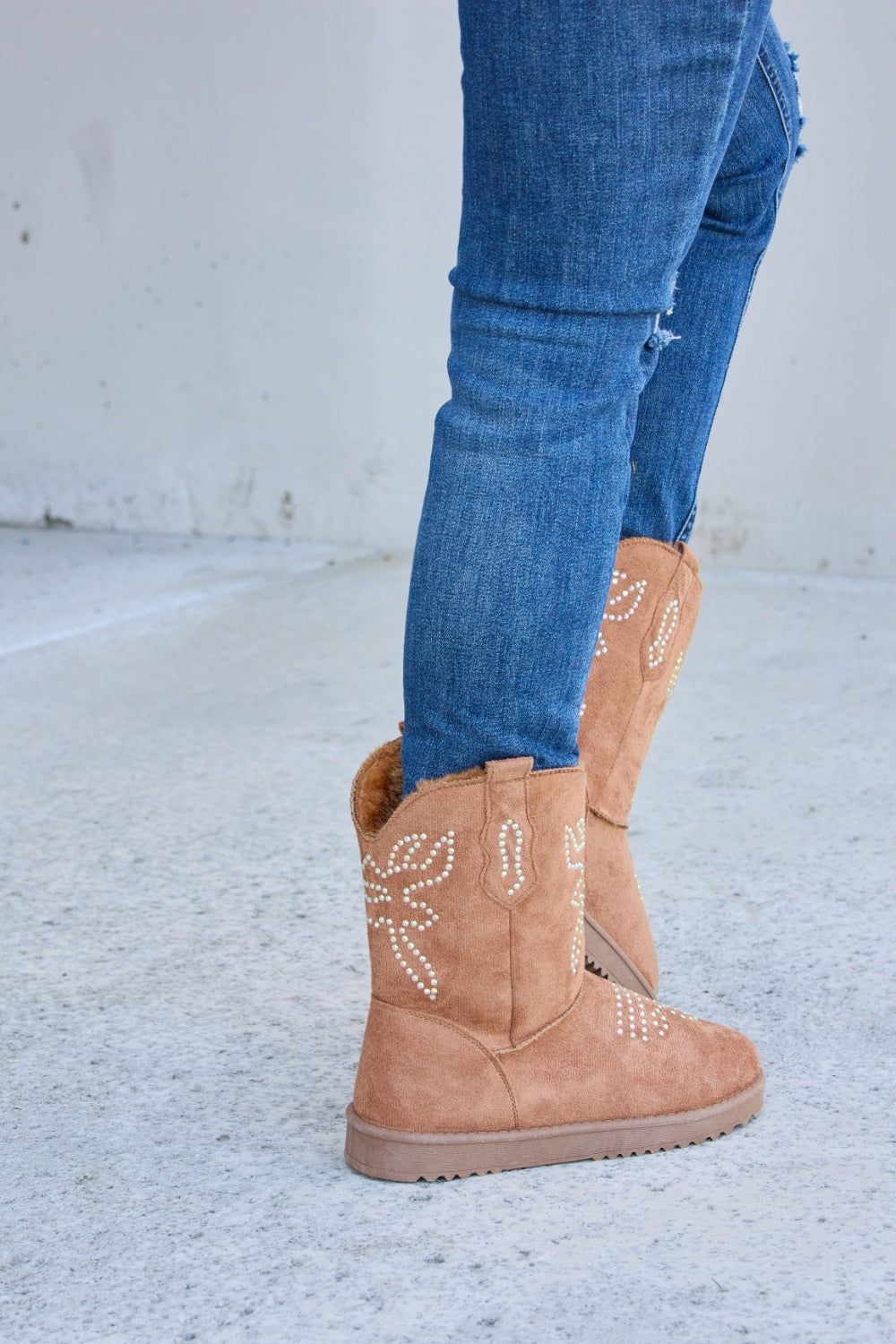 A person wearing ripped blue jeans and Forever Link Rhinestone Furry Flat Boots stands on a concrete surface.