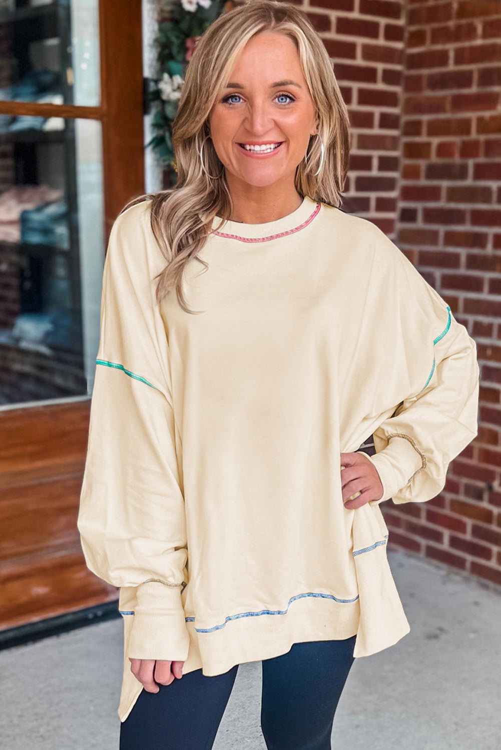 A woman with blonde hair is facing away, wearing a Contrast Stitching Split Baggy Sweatshirt. She stands in a room adorned with plants and wooden furniture in the background.