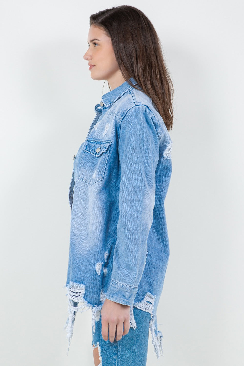 A woman stands against a plain background wearing the American Bazi Raw Hem Distressed Long Sleeve Denim Jacket over a white crop top and ripped jeans, adding an edgy flair to her wardrobe.