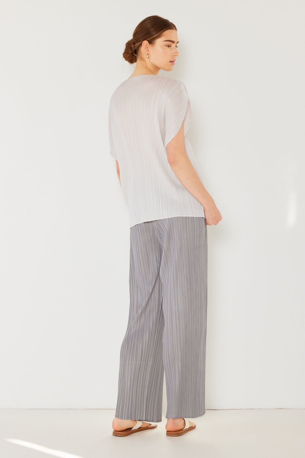 A person stands against a plain white background, wearing a sleeveless black top and versatile and stylish Marina West Swim Pleated Elastic-Waist Straight Pants, complemented by white sandals. They have light skin and brown hair tied back, and their arms are at their sides, highlighting the straight-leg design of the pants.