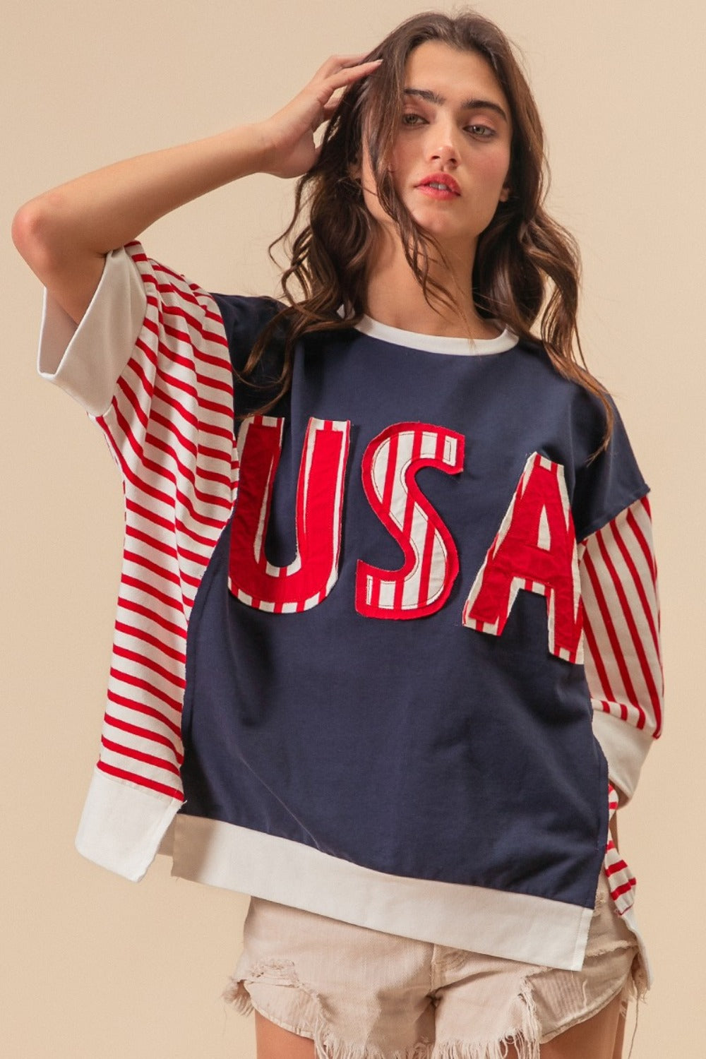 A person dressed in a “BiBi USA Letter Patchwork Contrast Short Sleeve T-Shirt” featuring blue, red, and white colors with striped sleeves, paired with beige shorts, poses against a beige background, proudly displaying their American spirit.