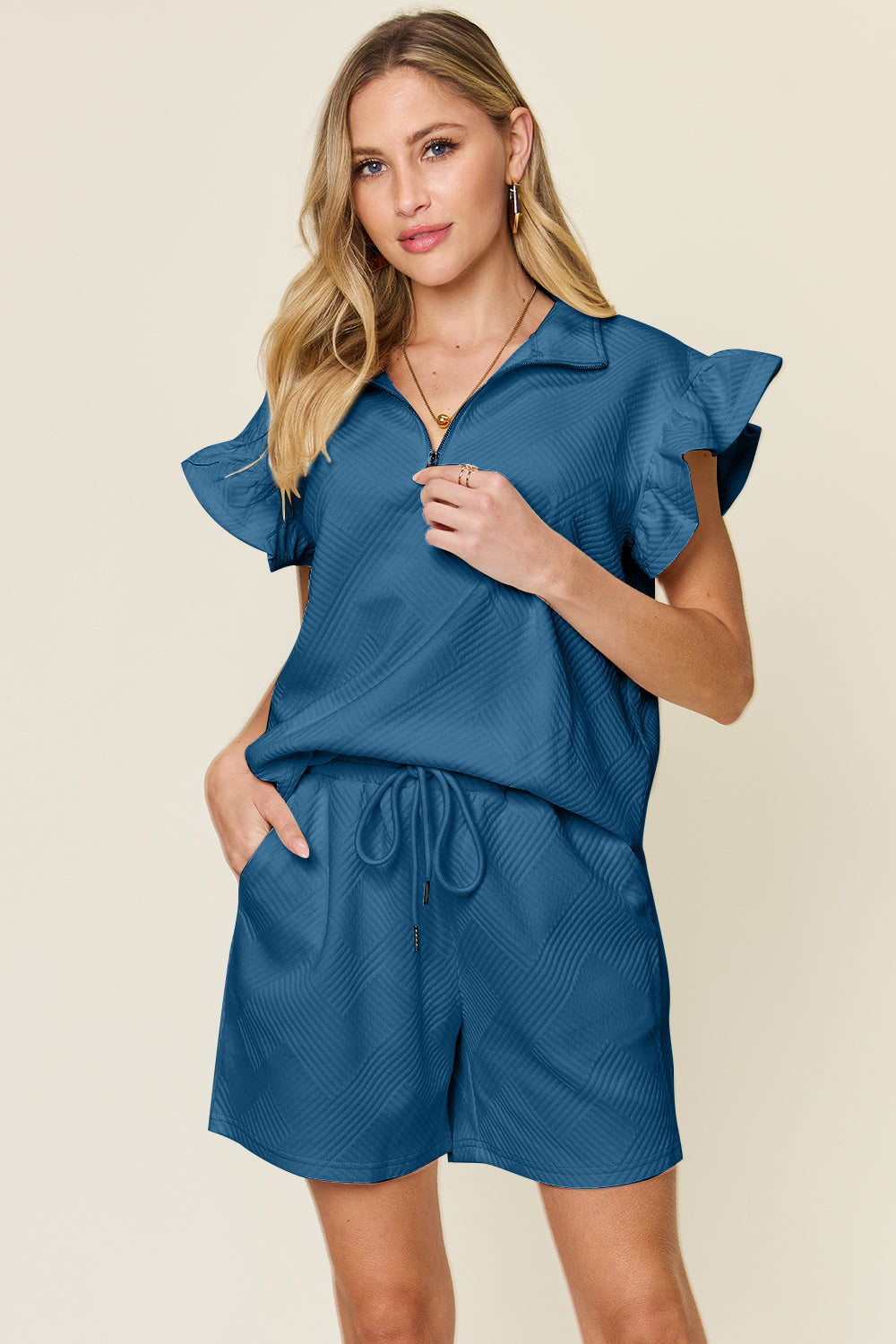 Against a plain background, a woman with blonde hair poses confidently with one hand in her pocket, highlighting the chic style of her Double Take Full Size Texture Flounce Sleeve Top and Drawstring Shorts Set.