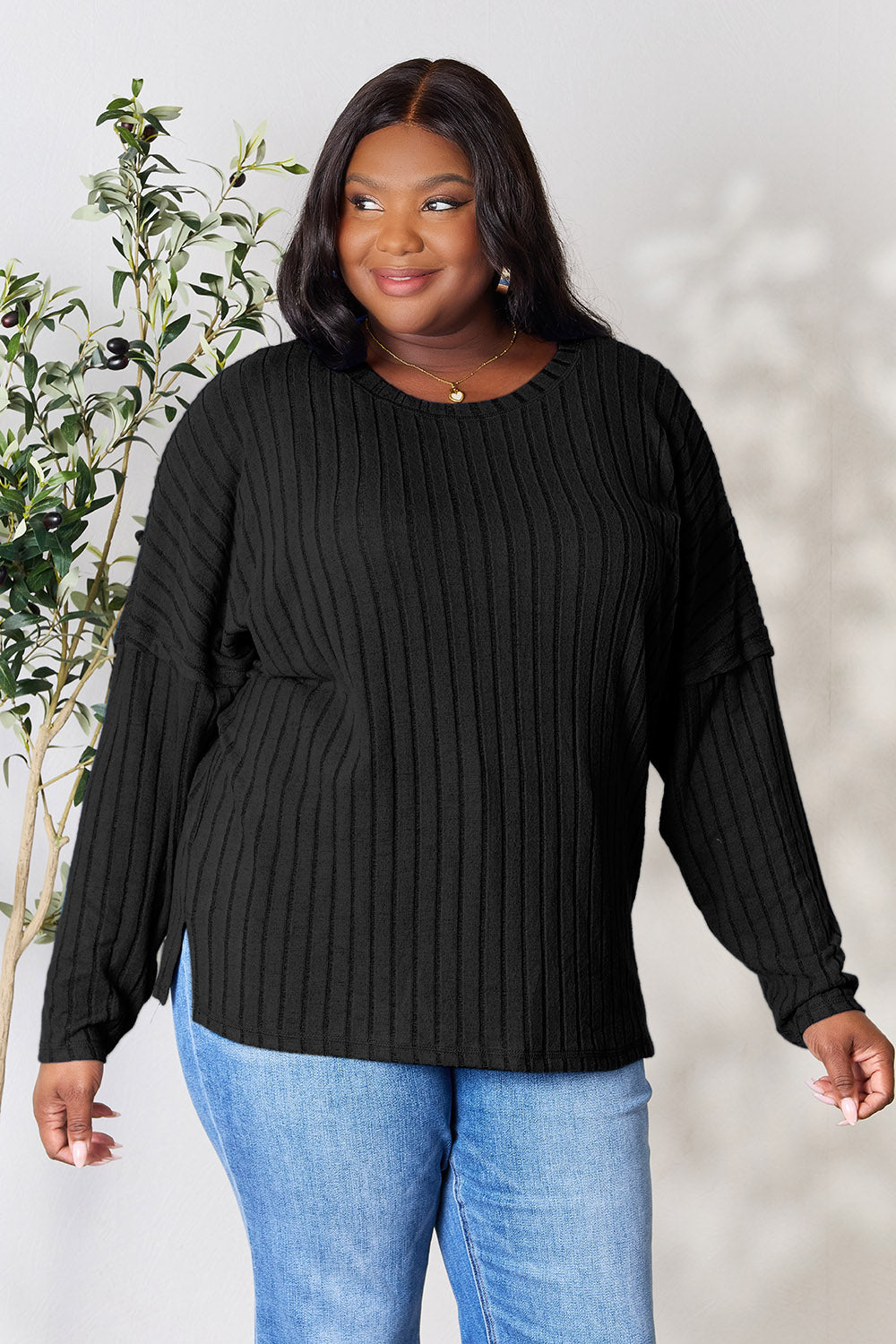 A person wearing the Basic Bae Full Size Ribbed Round Neck Slit Knit Top in blue and beige pants stands and smiles in front of a leafy plant.