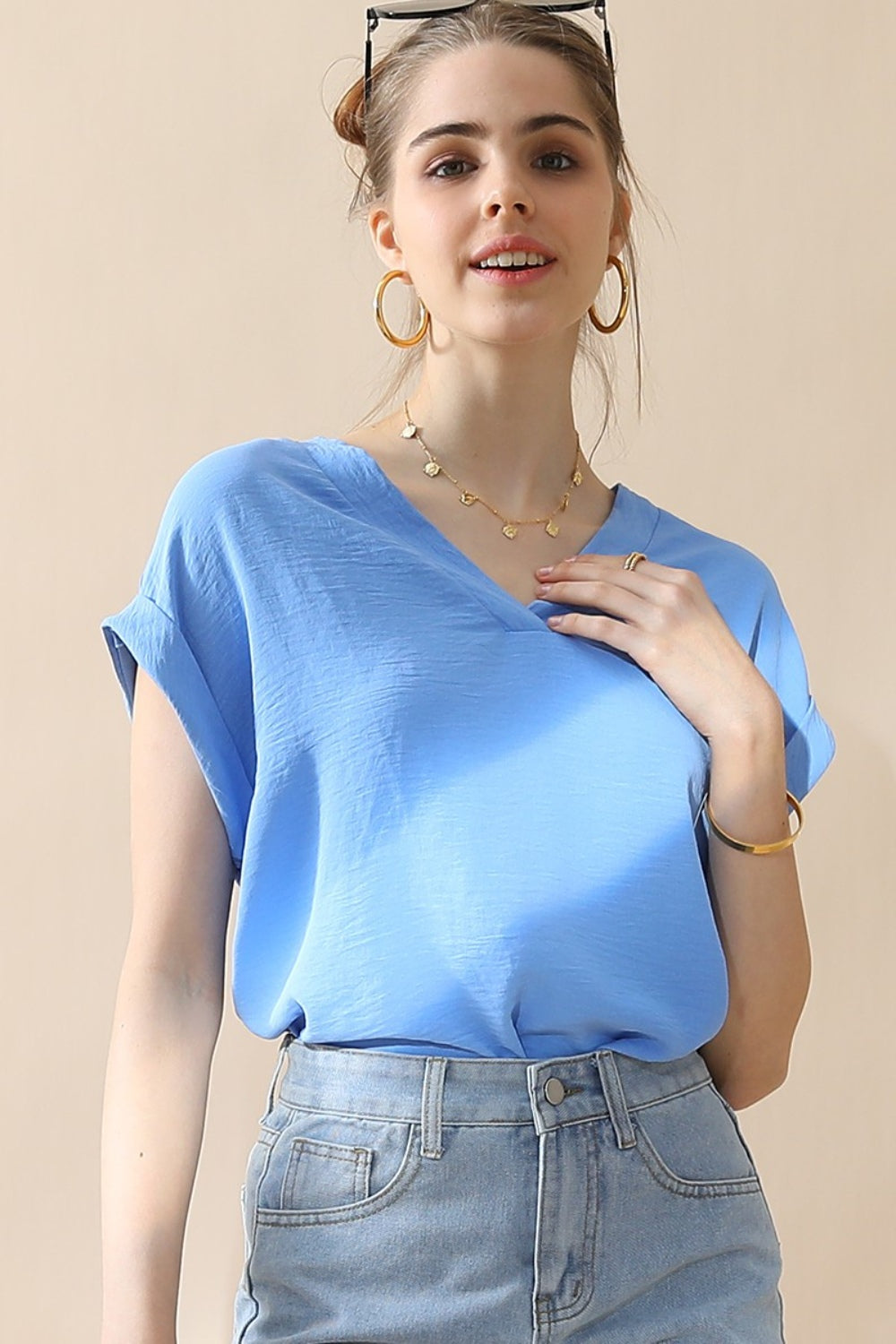 A woman wearing a stylish Ninexis V-Neck Trim Rolled Short Sleeve Shirt in light blue and high-waisted blue jeans, accessorized with hoop earrings, a necklace, and sunglasses on her head, poses in front of a beige background.