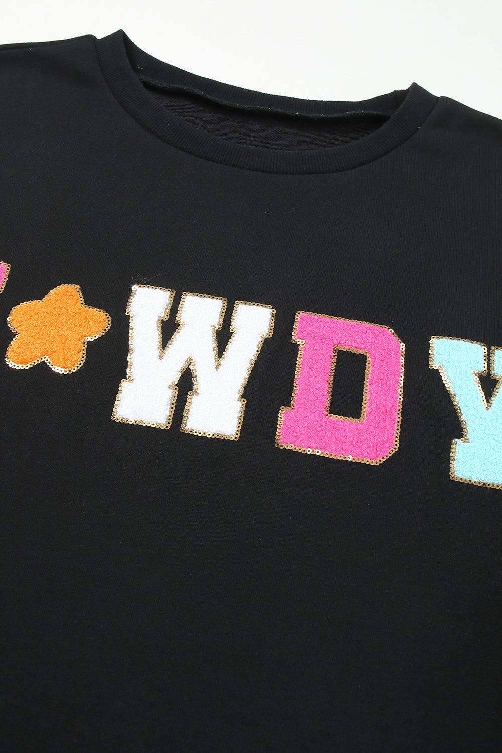 A woman with short blonde hair is wearing the Black Howdy Glitter Chenille Patch Graphic Casual Sweatshirt, featuring colorful star patterns and "WD" letters. She pairs it with light denim shorts and smiles while looking to the side.