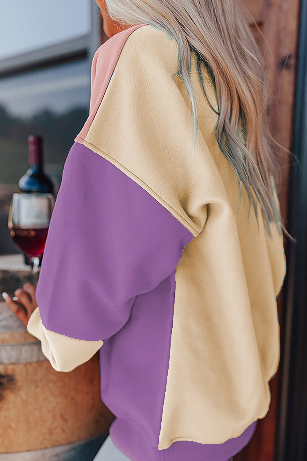 A woman with long blonde hair stands with her back to the camera, wearing a trendy Meadow Mauve Colorblock Patchwork Drop Shoulder Sweatshirt. She is next to a wooden barrel and large pampas grass decorations.