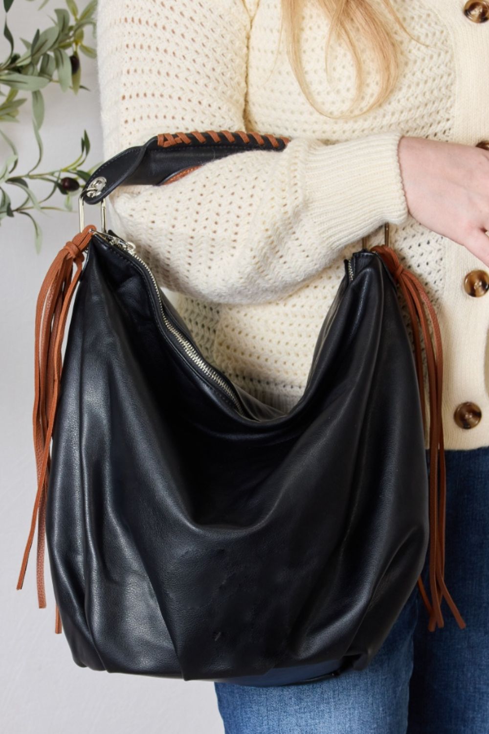 Individual holding a SHOMICO Fringe Detail Contrast Handbag, featuring cream and brown accents, paired with a knitted cream sweater, standing against a light background. The handbag exudes Bohemian flair with its relaxed style.