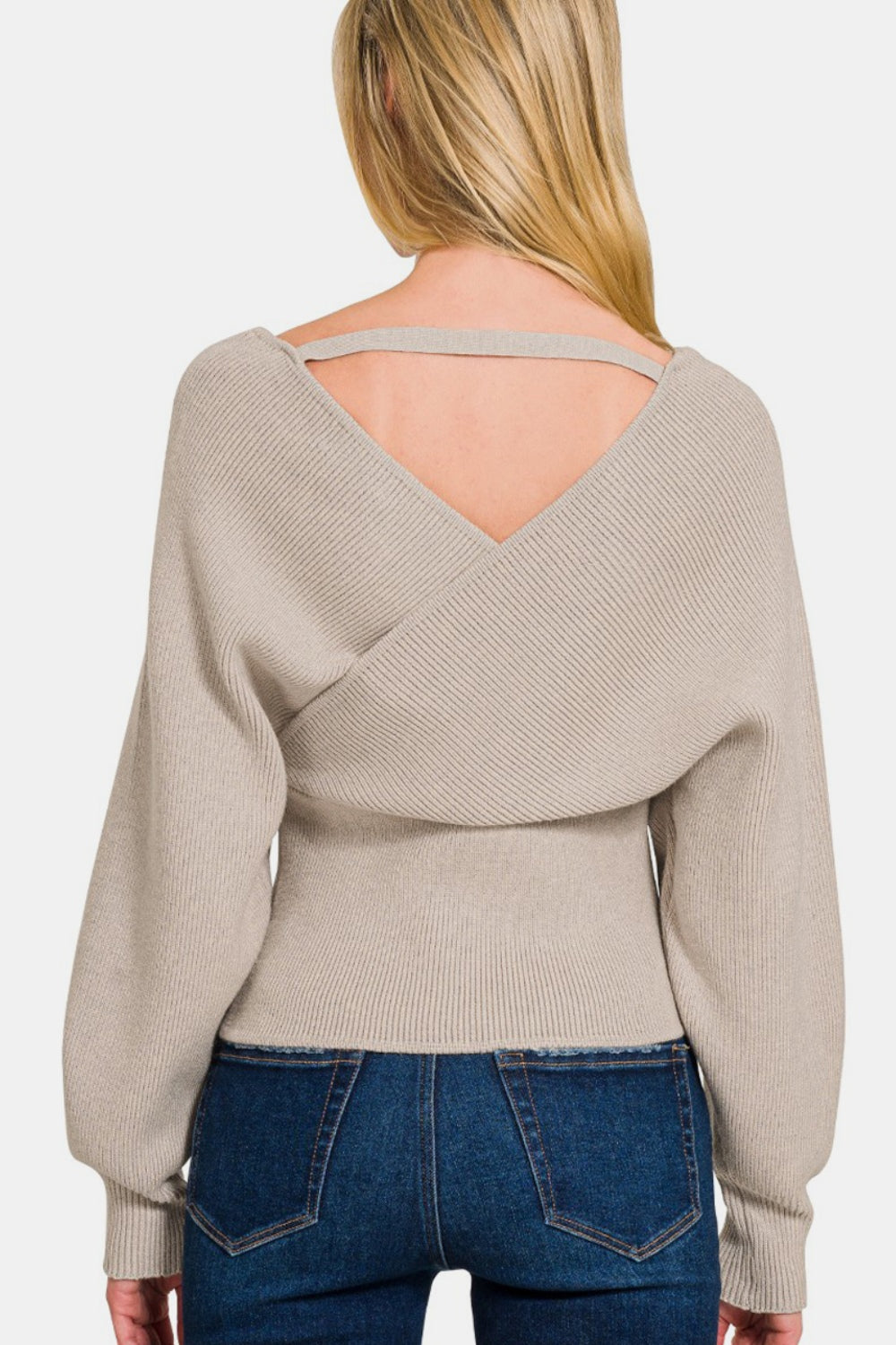 A woman with long blonde hair is smiling with one hand in her pocket against a plain white background, wearing the Zenana Cross Wrap Rib Long Sleeve Sweater in beige and blue jeans—a perfect layering option for any season.