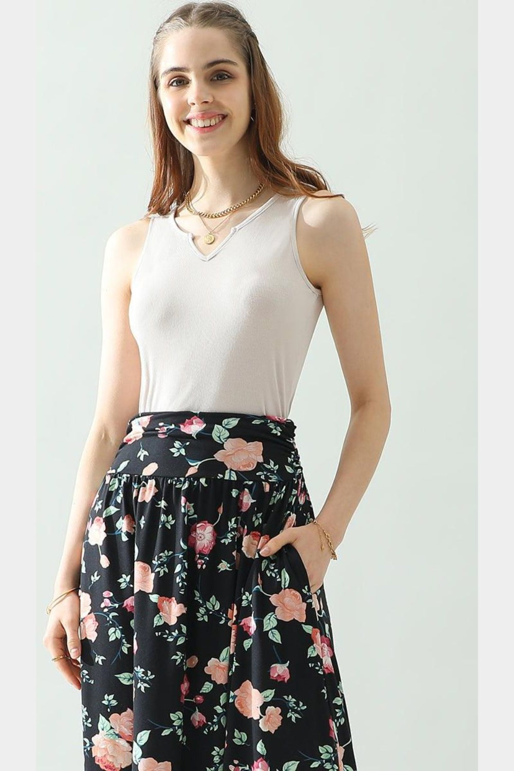 A woman wearing the Ninexis Notched Rib Knit Tank in white paired with a floral skirt and accessorized with a necklace, standing against a light background. This chic look is perfect for any summer wardrobe.