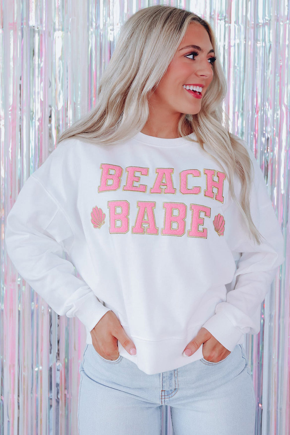 A person with long blonde hair is standing with their back to the camera, wearing a White BEACH BABE Slogan Graphic Casual Sweatshirt and light blue jeans.