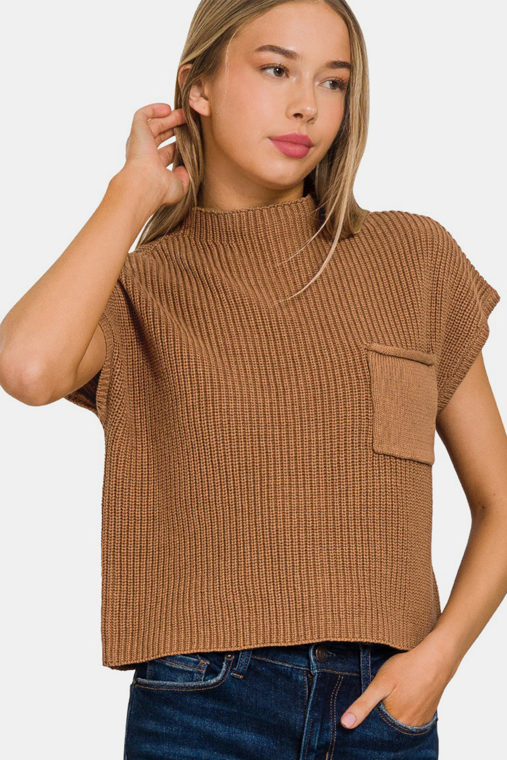 A person with long hair is wearing a Zenana Mock Neck Short Sleeve Cropped Sweater in brown and blue jeans. They are touching their hair with one hand and looking to the side, showcasing the casual comfort of their attire.