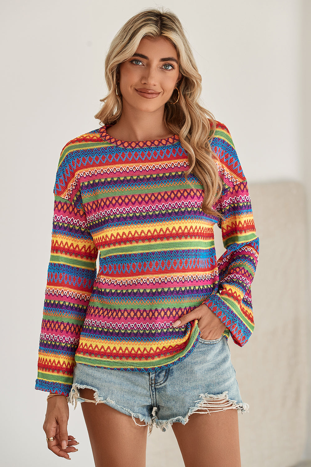 A person stands with hands in the pockets of their denim shorts, wearing a colorful Rose Red Ethnic Wavy Pattern Drop Shoulder Knit Top and a white shoulder bag.