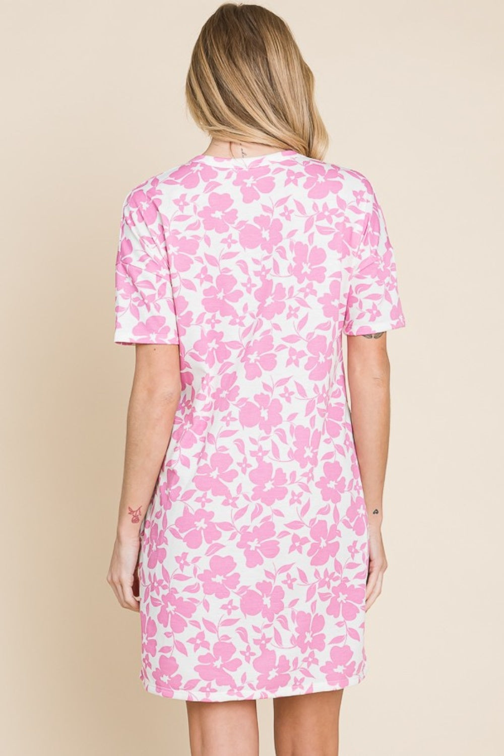 A person in a BOMBOM Floral Button Down Mini Dress, featuring a charming pink and white pattern, stands against a plain background, highlighting its versatile design and feminine silhouette.