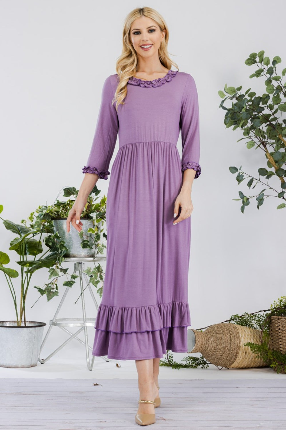 A woman in the Celeste Full Size Layered Ruffle Hem Dress with Pockets, featuring an A-line silhouette and 3/4 sleeves, stands on a wooden floor surrounded by potted plants and a wicker basket.