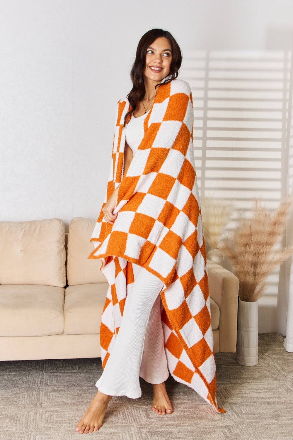 A person stands holding a large Cuddley Checkered Decorative Throw Blanket, smiling, in a room with a sofa and a potted plant. The blanket features a black and white checkerboard pattern, is made from 100% polyester, and imported for quality.