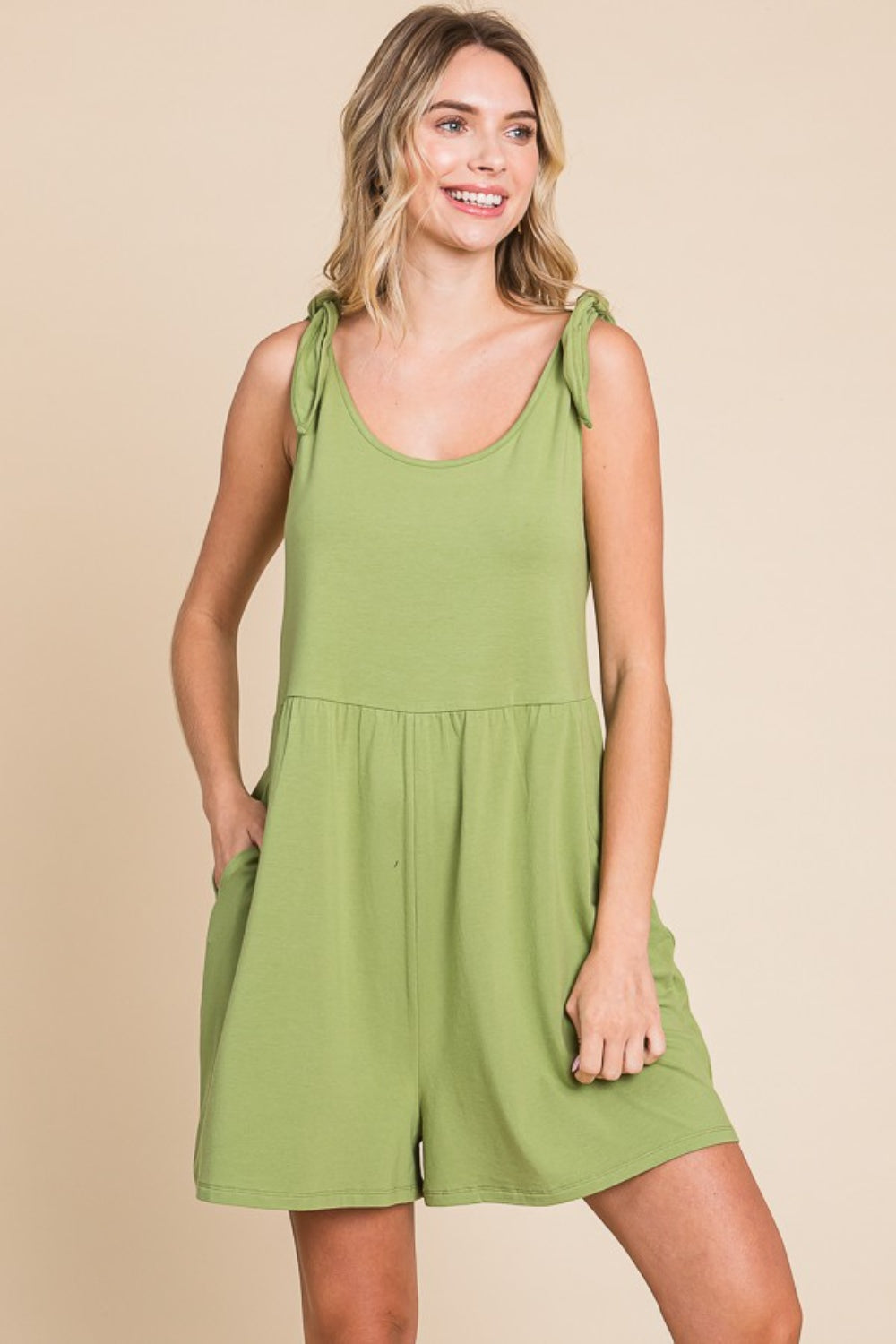 A person stands in the Culture Code Full Size Shoulder Knot Baggy Romper, featuring a sleeveless design and light green color composed of lightweight breathable fabric. With hands comfortably in pockets, they smile at the camera against a plain beige background.