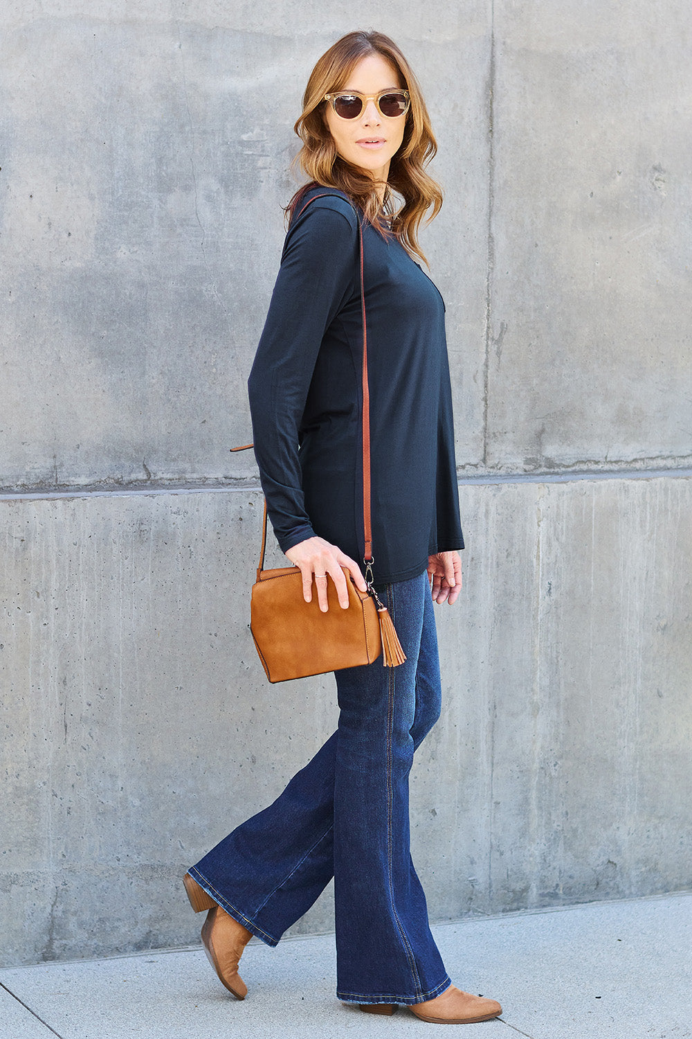 A woman with long hair wearing sunglasses, a Basic Bae Full Size Round Neck Long Sleeve Top in turquoise, and jeans is standing in front of a concrete wall with one hand on her hip.