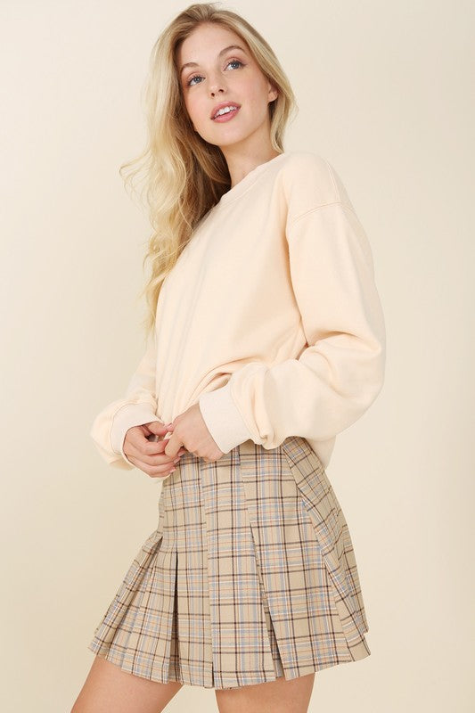 A person dressed in a plaid pleated mini skirt paired with a beige sweater is posed against a plain backdrop. This fashionable and versatile outfit, featuring the Plaid Pleated Mini Skirt, is designed for easy maintenance and can be machine washed in cold water.