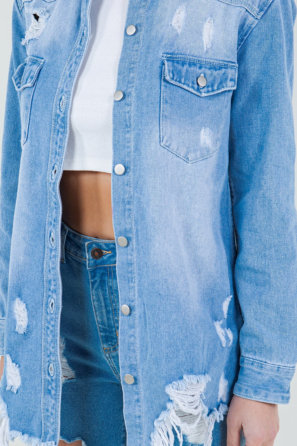 A woman stands against a plain background wearing the American Bazi Raw Hem Distressed Long Sleeve Denim Jacket over a white crop top and ripped jeans, adding an edgy flair to her wardrobe.