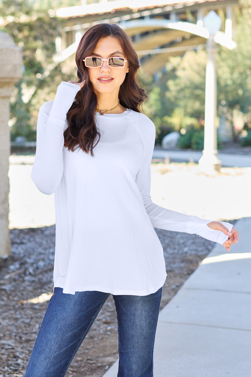 A woman wearing sunglasses and a slightly stretchy Basic Bae Full Size Round Neck Long Sleeve T-Shirt in white stands on a sidewalk with an arched structure in the background, smiling slightly.