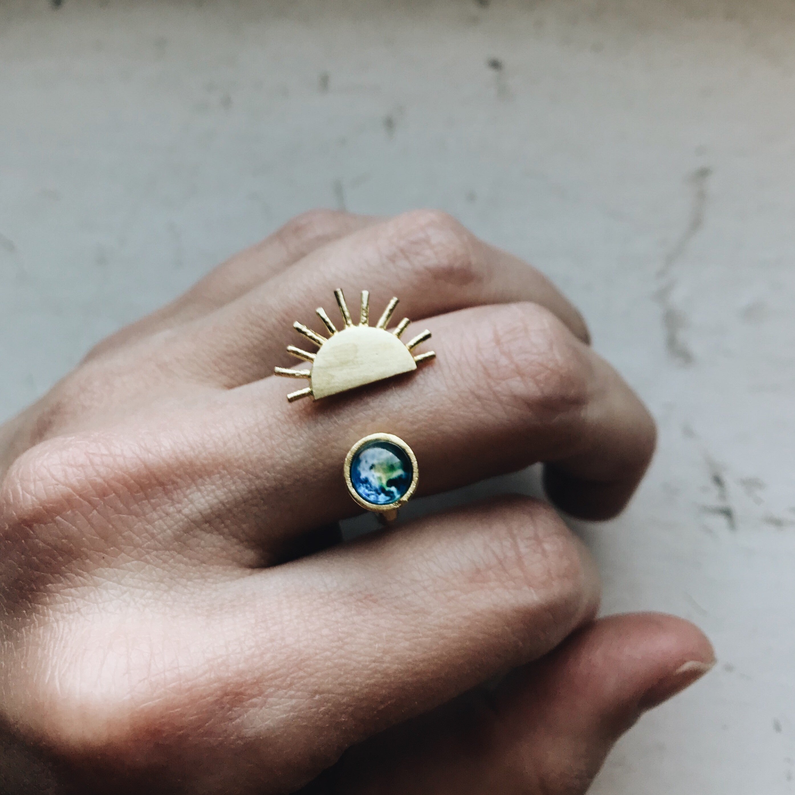 A hand showcases two exquisite pieces of Sunrise jewelry: a gold statement ring featuring a sun design and another adorned with a small globe image, both capturing the essence of elegance and adventure.