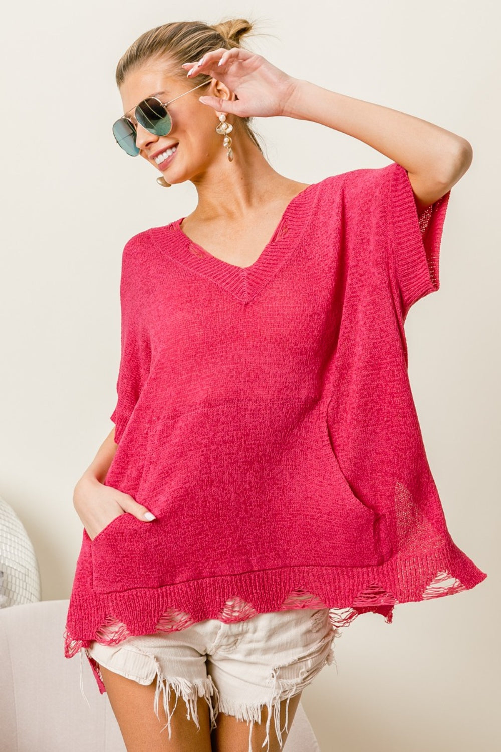 A woman in sunglasses, wearing a BiBi Distressed Hem V-Neck Slit Sweater in red and white shorts, smiles with her hands in her pockets, effortlessly showcasing her modern edge fashion.